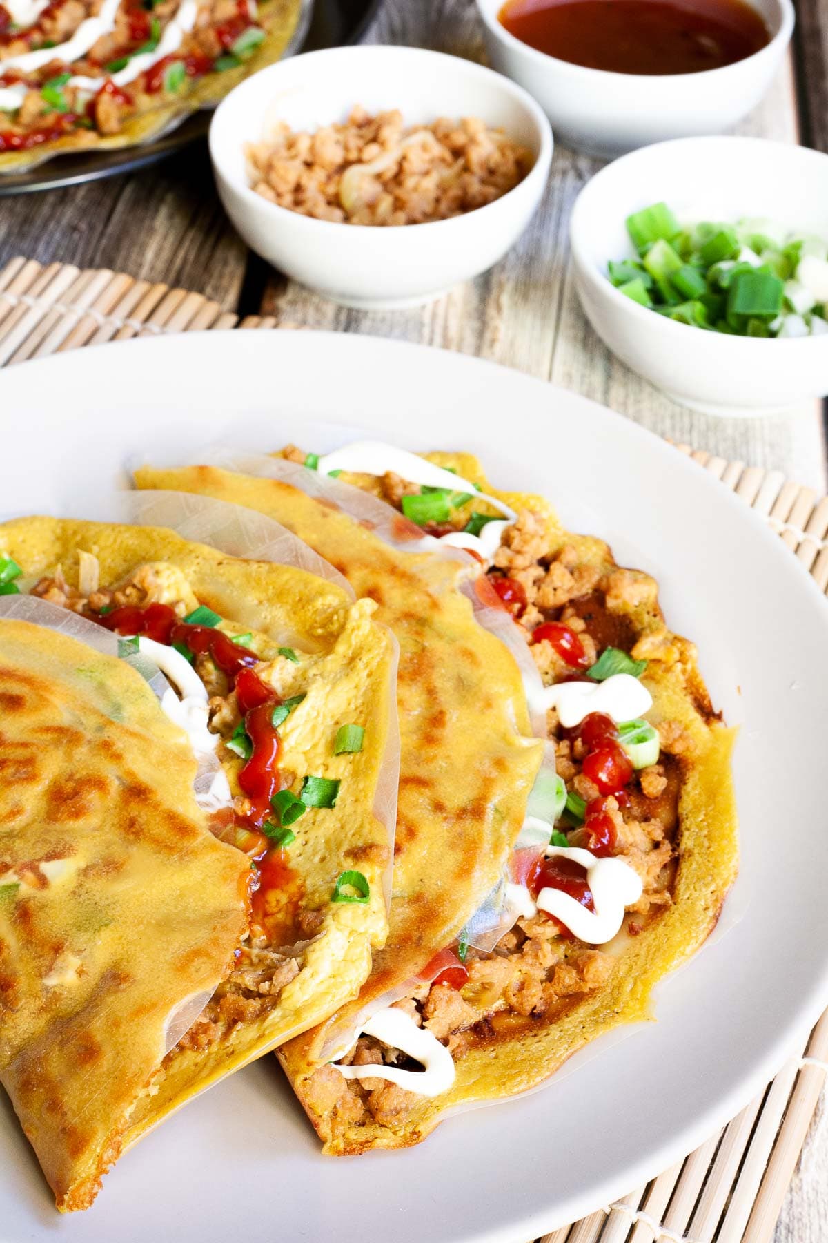 White plate with 2 folded rice paper pizzas topped with brown crumbles, green onion rings, and drizzled with white and red sauces