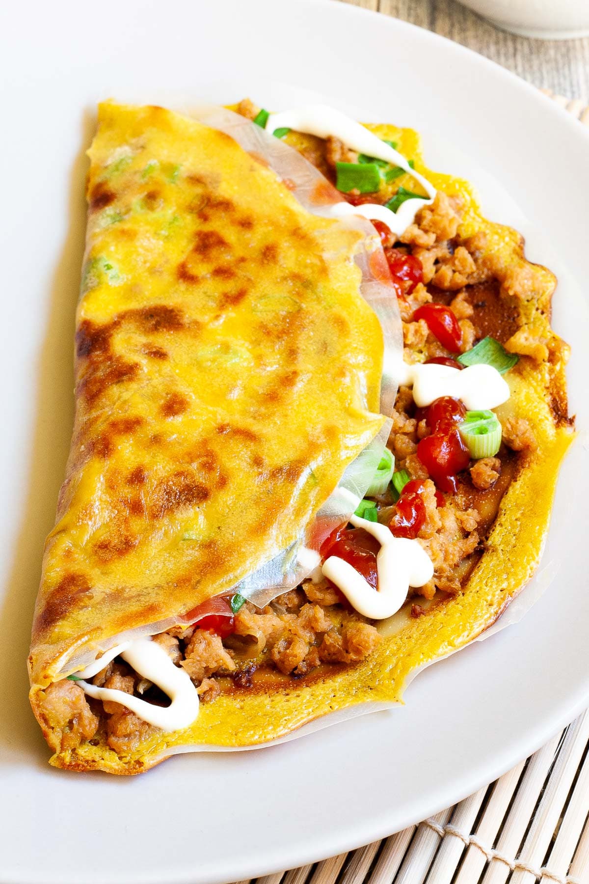 White plate with a folded rice paper pizza topped with brown crumbles, green onion rings, and drizzled with white and red sauces