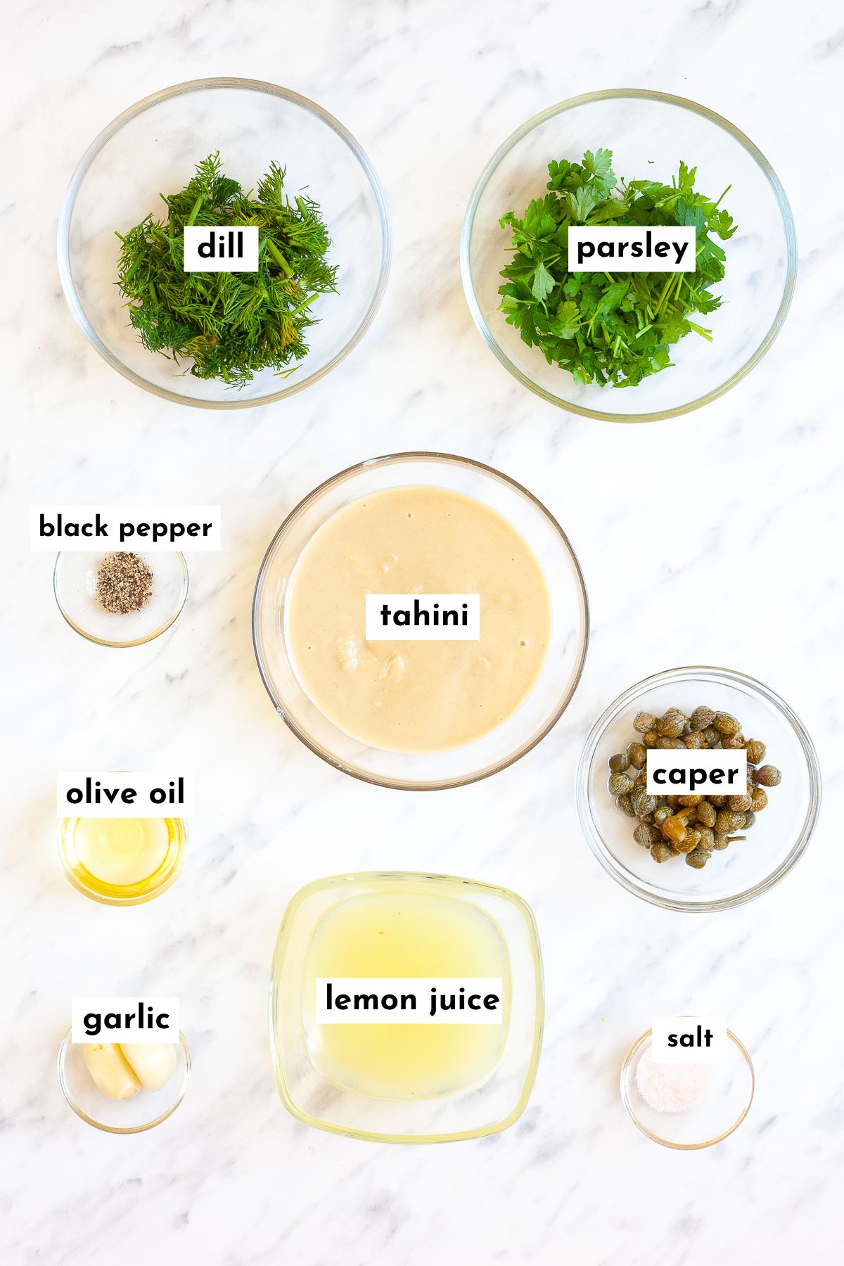 Ingredients of lemon herb tahini sauce is small glass bowls dill, parsley, tahini, lemon juice, capers, salt, pepper, olive oil and garlic powder