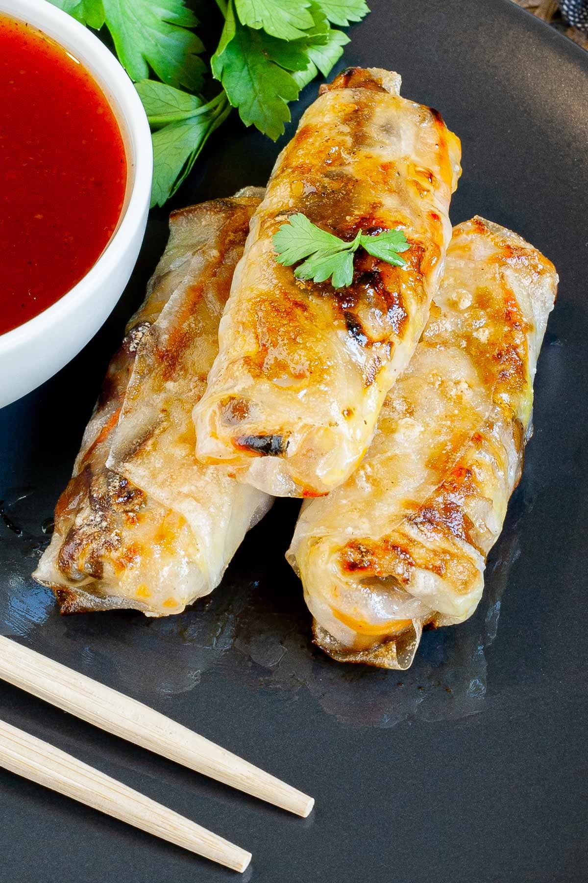 Black plate with crispy looking golden brown egg rolls. A small white bowl is with a red dipping sauce. The rolls are sprinkled with parsley leaves.