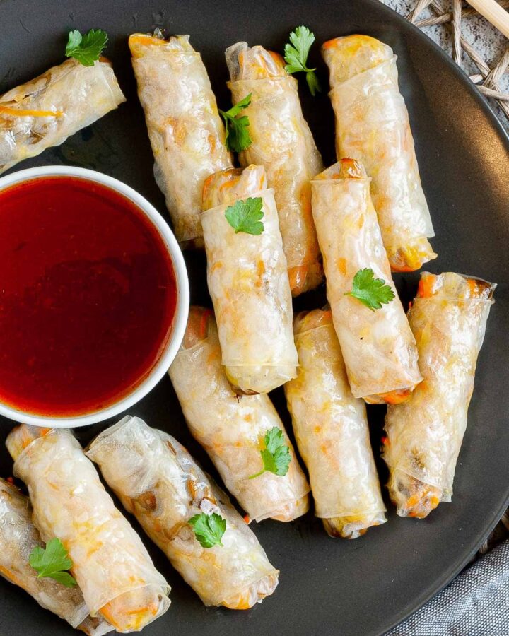 Black plate with crispy looking light brown egg rolls. A small white bowl is with a red dipping sauce. The rolls are sprinkled with parsley leaves.