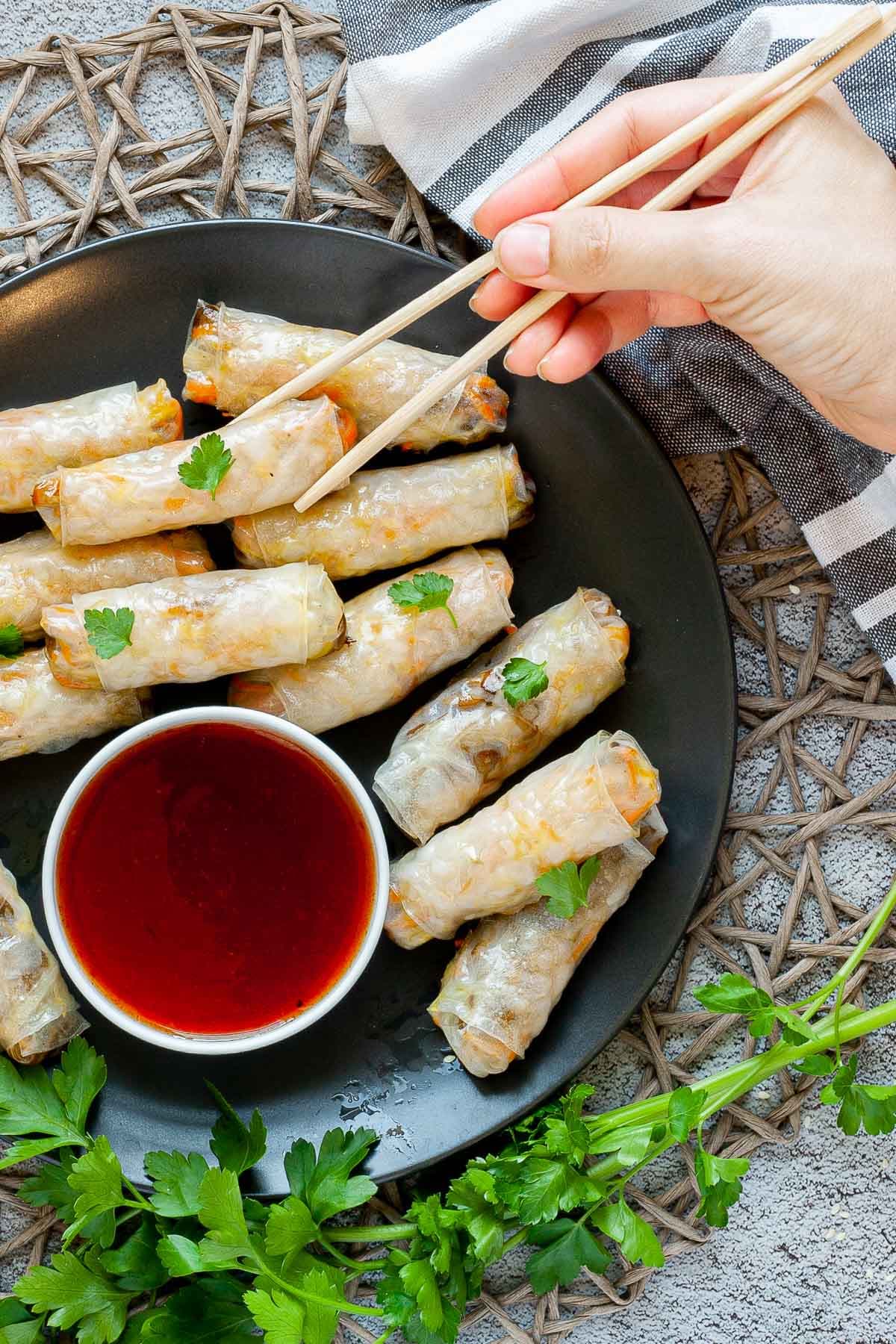 Black plate with crispy looking light brown egg rolls. A small white bowl is with a red dipping sauce. The rolls are sprinkled with parsley leaves.