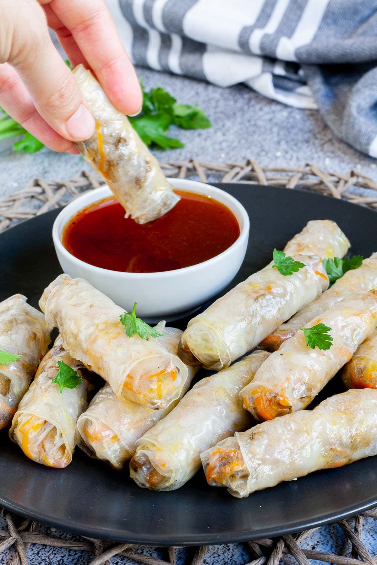 Black plate with crispy looking light brown egg rolls. A small white bowl is with a red dipping sauce. The rolls are sprinkled with parsley leaves. A hand is dipping one roll in the sauce.