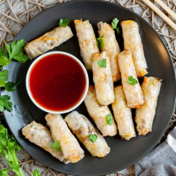 Black plate with crispy looking light brown egg rolls. A small white bowl is with a red dipping sauce. The rolls are sprinkled with parsley leaves.