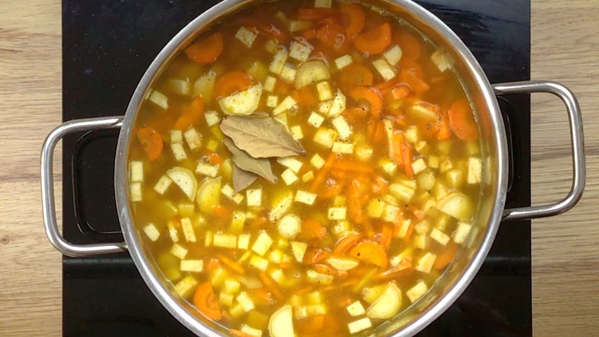 Large pot with a light brown sauce, bay leaves, and lots of chopped orange and white veggies like carrot, celeriac and parsnip.