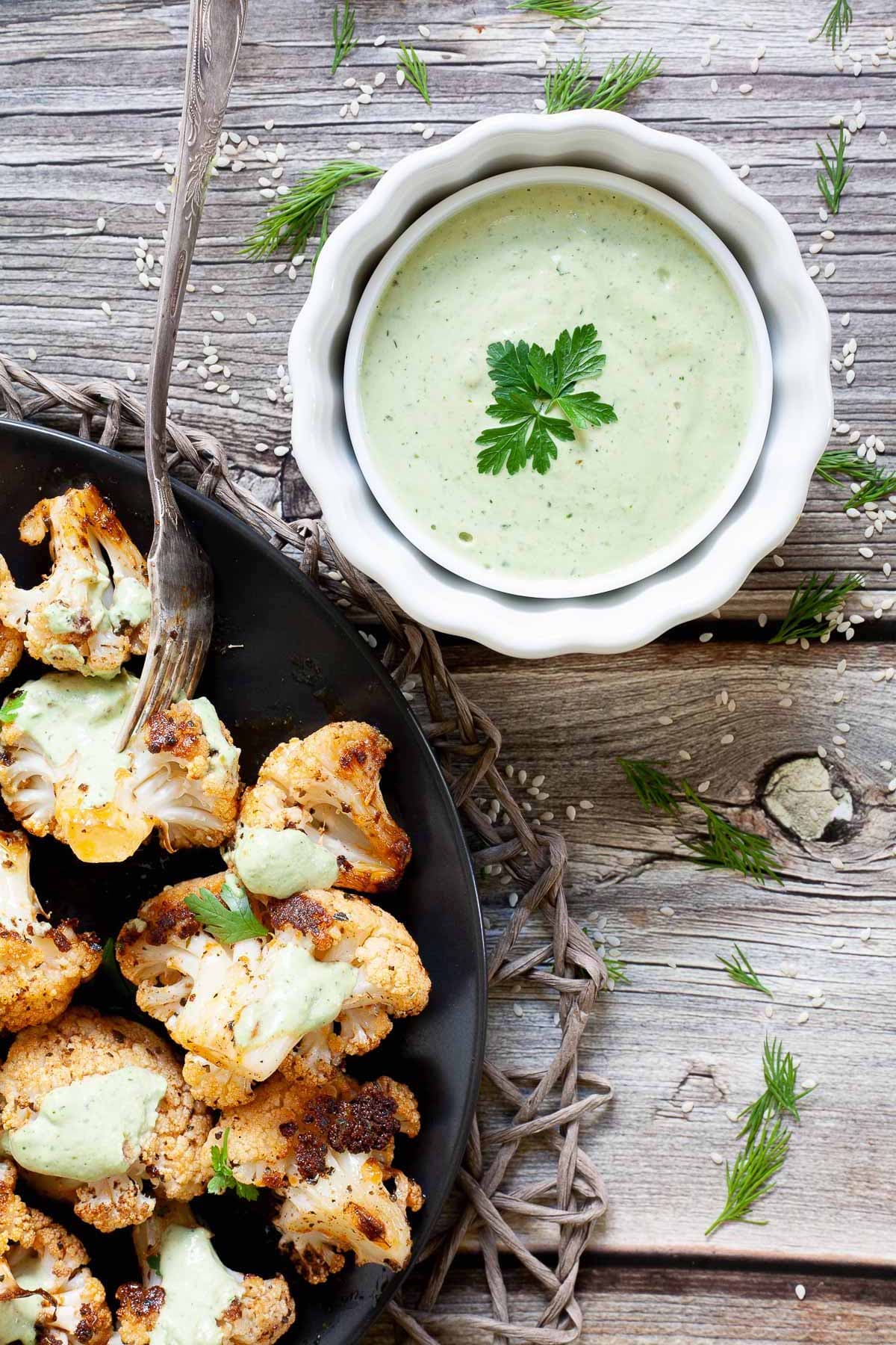 Black plate full of roasted cauliflower florets drizzled with a light green creamy sauce and sprinkled with fresh parsley is at the corner. And a small white bowl with the green creamy sauce is in the middle.