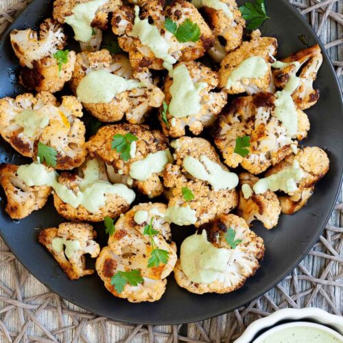 Black plate full of roasted cauliflower florets drizzled with a light green creamy sauce and sprinkled with fresh parsley. a small white bowl with the green creamy sauce is next to it.