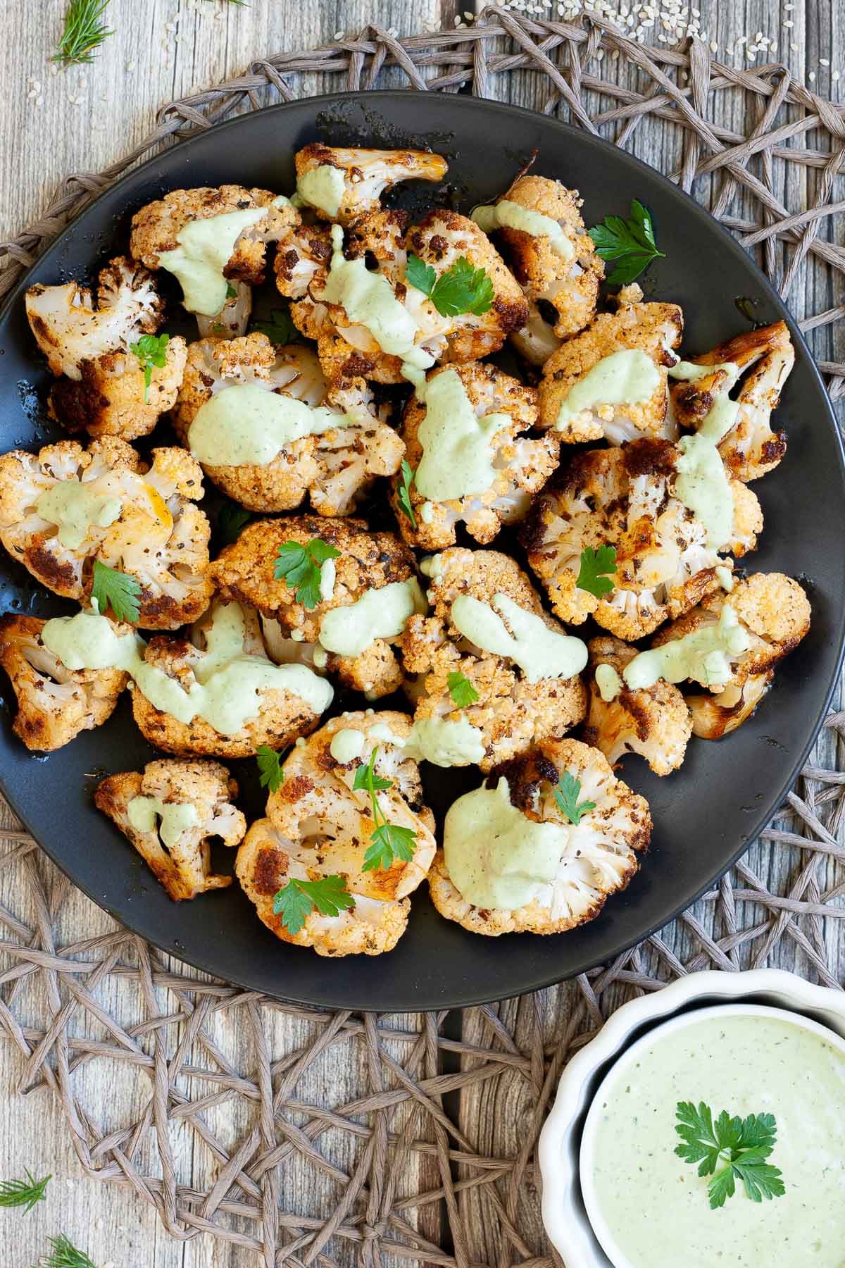 Black plate full of roasted cauliflower florets drizzled with a light green creamy sauce and sprinkled with fresh parsley. a small white bowl with the green creamy sauce is next to it. 