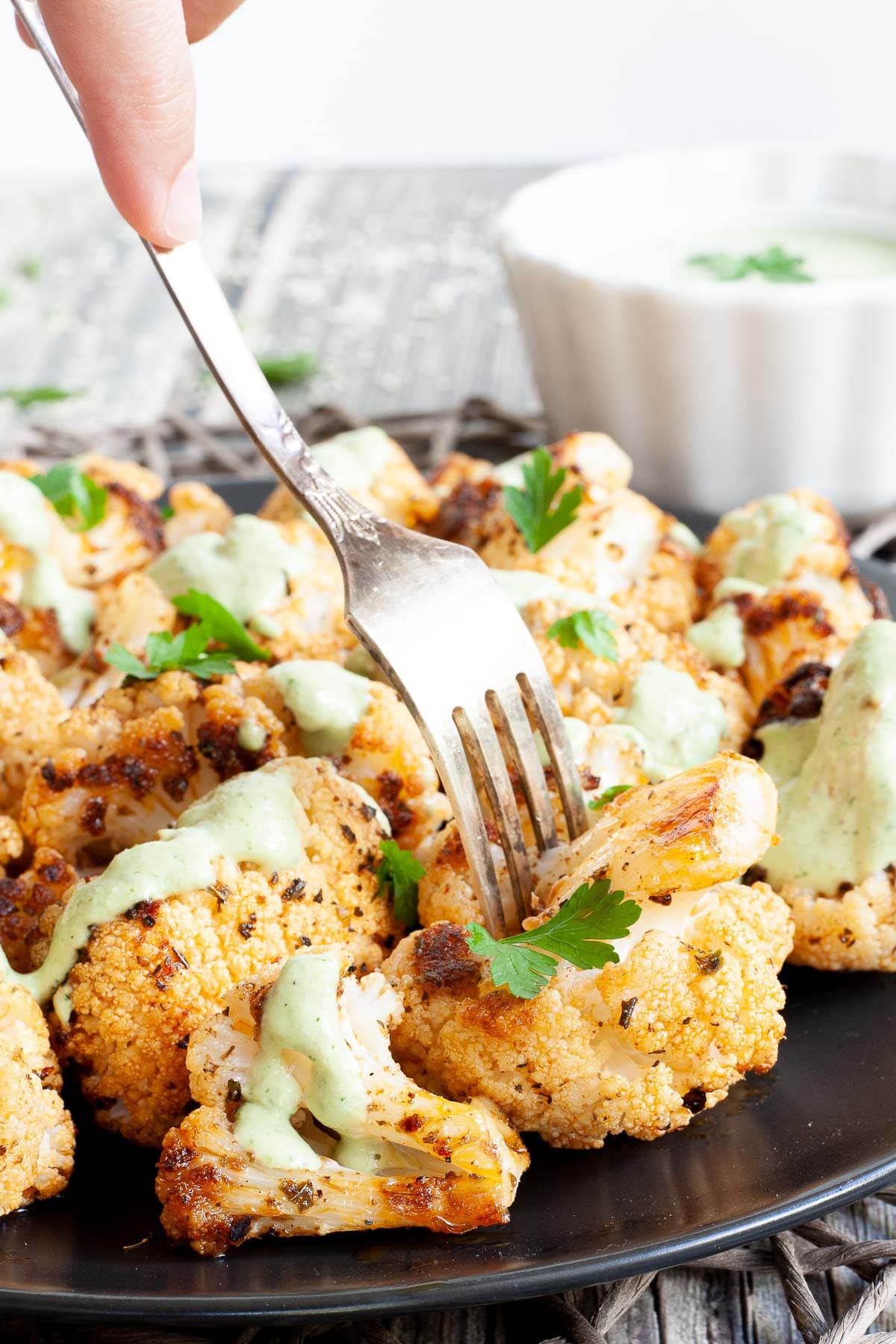 Black plate full of roasted cauliflower florets drizzled with a light green creamy sauce and sprinkled with fresh parsley. A hand is holding a fork and taking one floret.