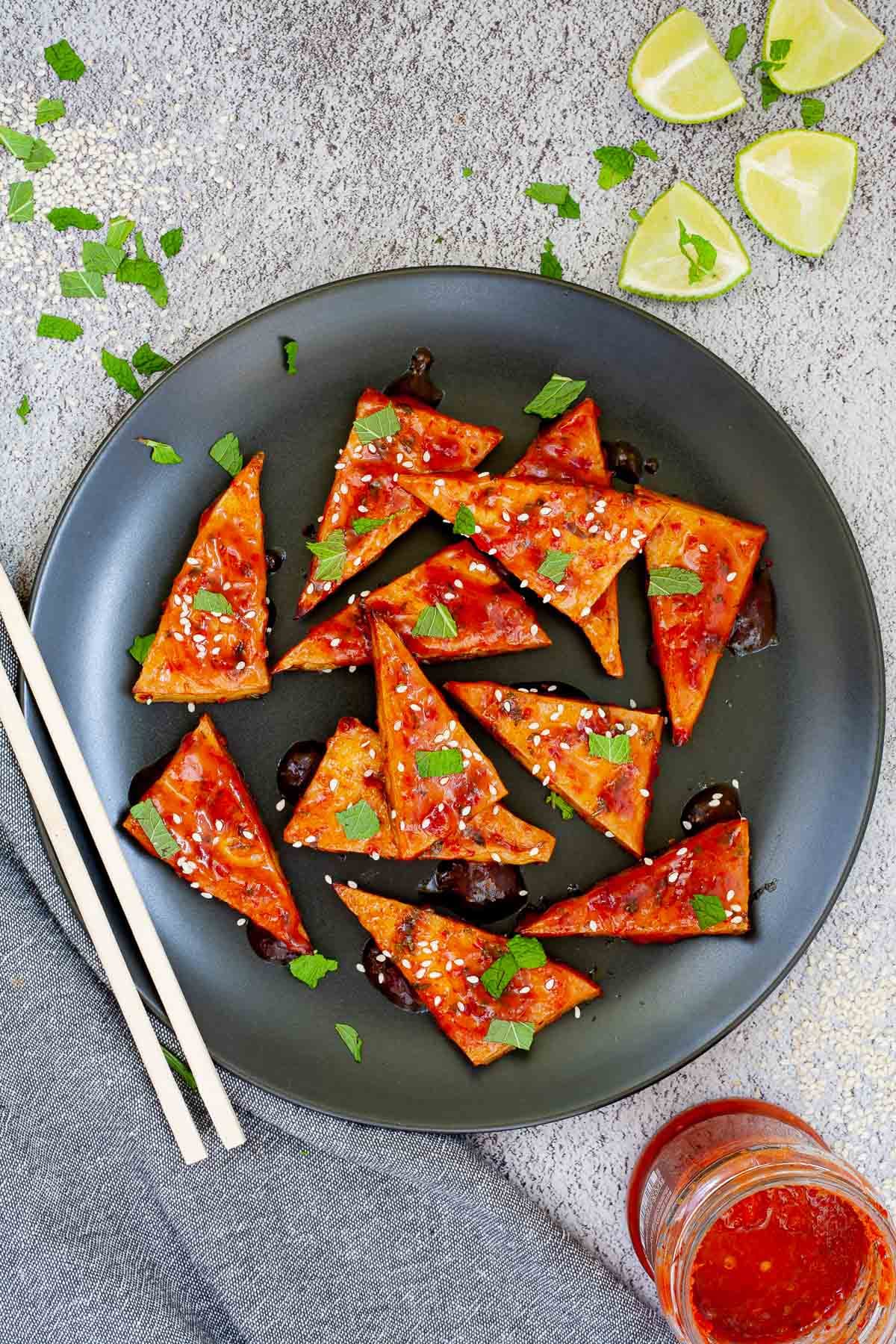 Triangle-shaped tofu slices in a red sticky glaze with freshly chopped green mint, sesame seeds on black plate