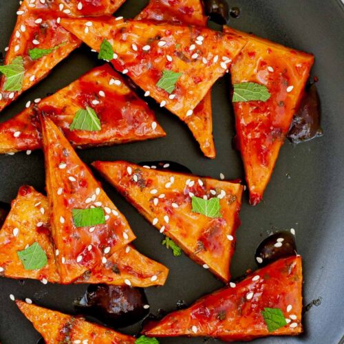Triangle-shaped tofu slices in a red sticky glaze with freshly chopped green mint, sesame seeds on black plate