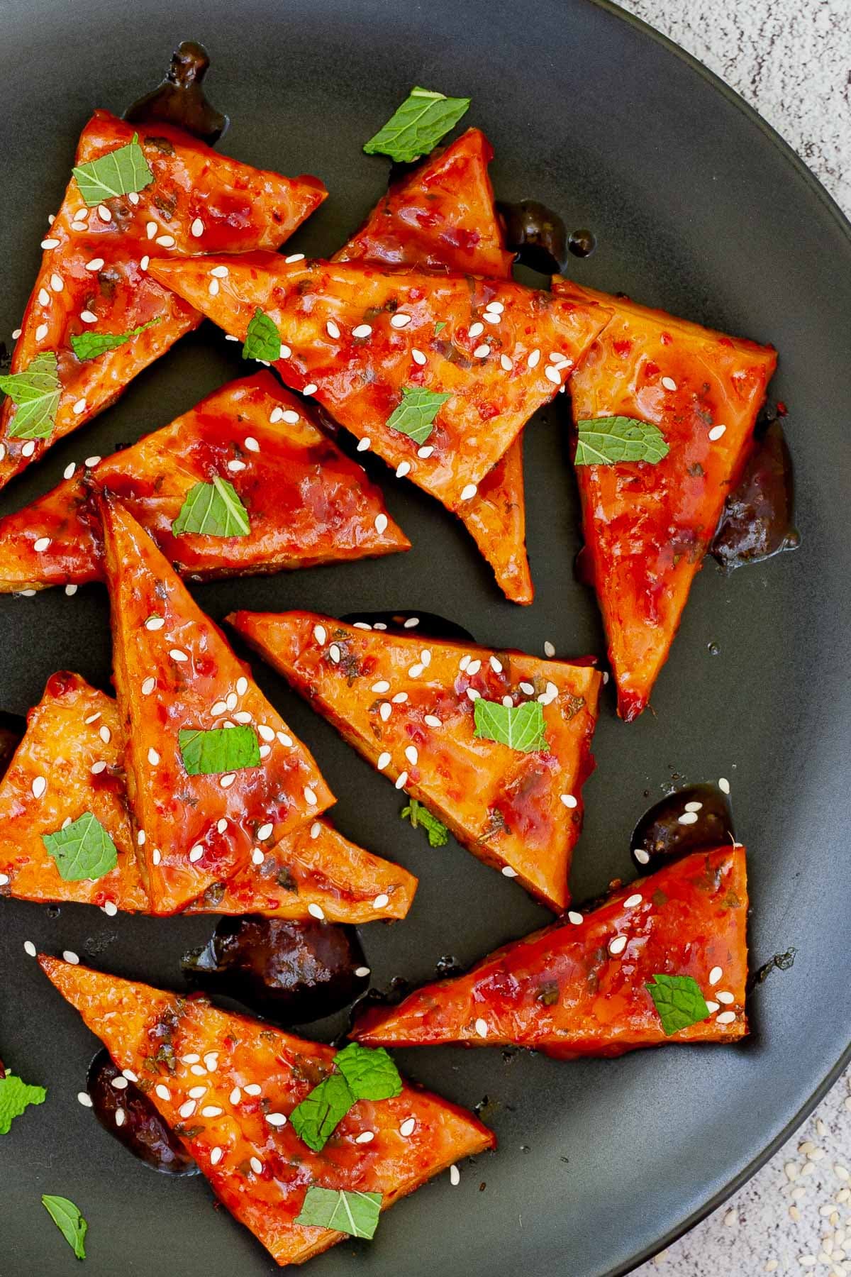 Triangle-shaped tofu slices in a red sticky glaze with freshly chopped green mint, sesame seeds on black plate