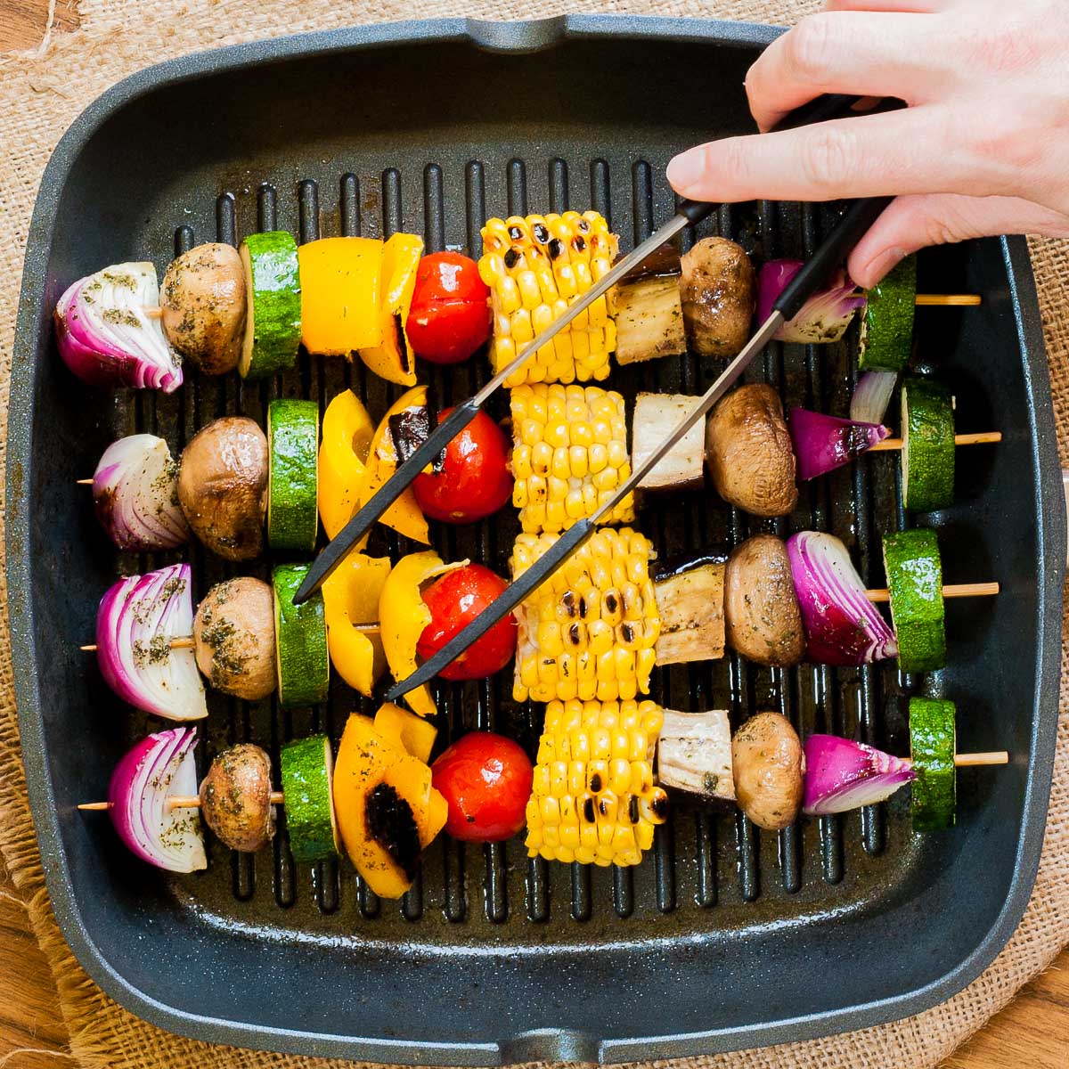 Grill pan with 4 veggie kabobs onion, mushroom, zucchini, bell pepper, cherry tomato, corn, eggplant slices. A hand is holding tongs to flip them.