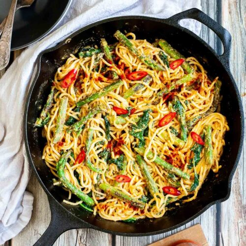 Black cast iron skillet with spaghetti in creamy light brown sauce with asparagus, spinach and red pepper slices. Red harissa sauce in a glass jar and a spoon with tahini paste next to it.