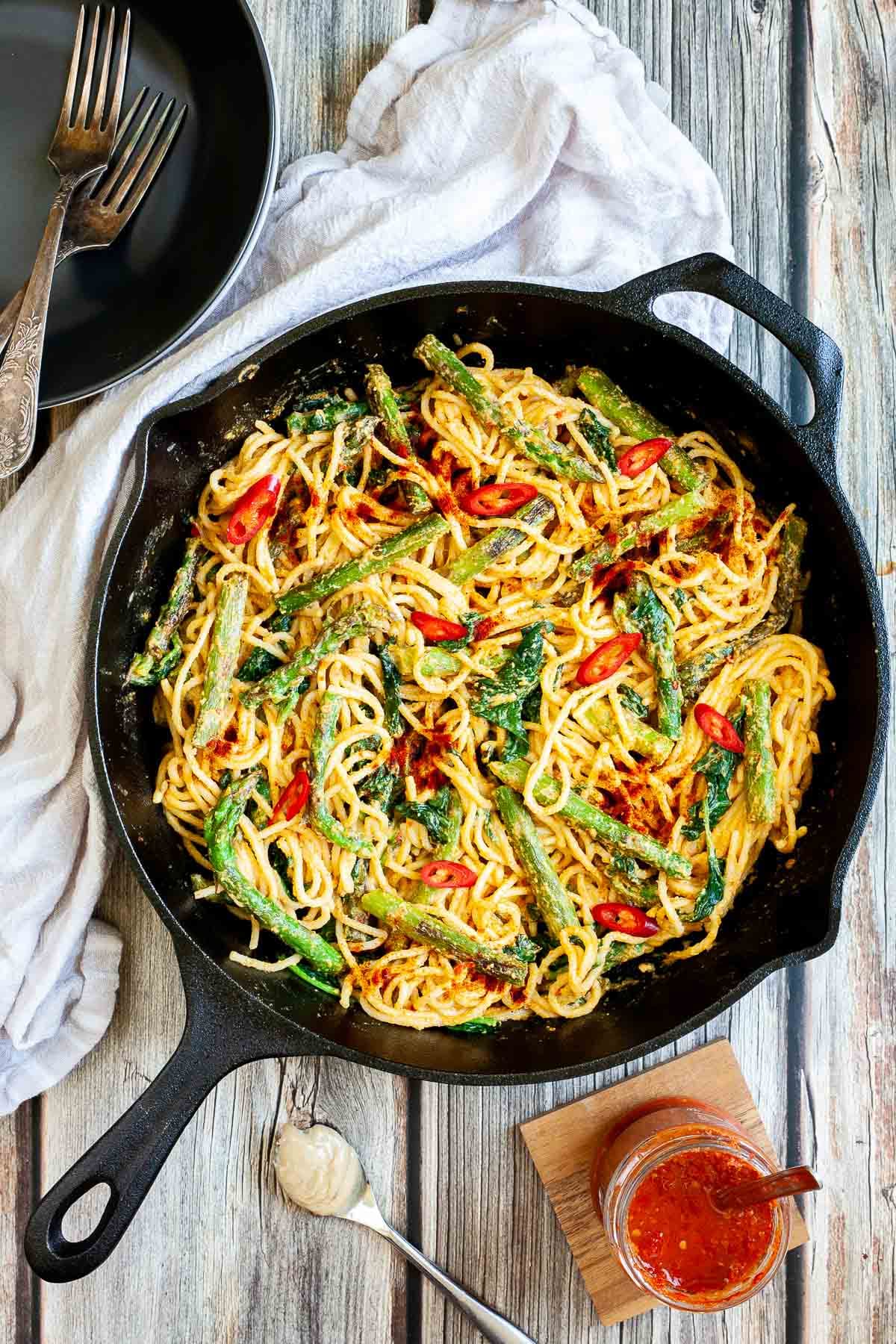 Black cast iron skillet with spaghetti in creamy light brown sauce with asparagus, spinach and red pepper slices. Red harissa sauce in a glass jar and a spoon with tahini paste next to it.