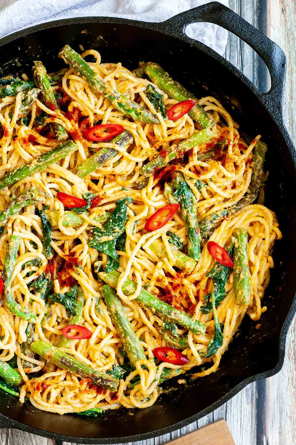 Black cast iron skillet with spaghetti in creamy light brown sauce with asparagus, spinach and red pepper slices