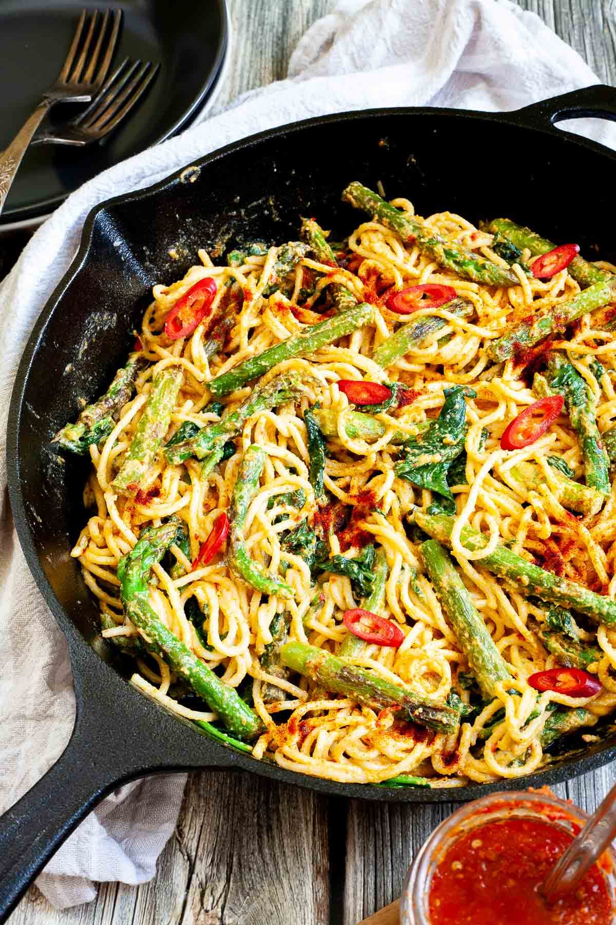 Black cast iron skillet with spaghetti in creamy light brown sauce with asparagus, spinach and red pepper slices. With harissa sauce in glass jar next to it