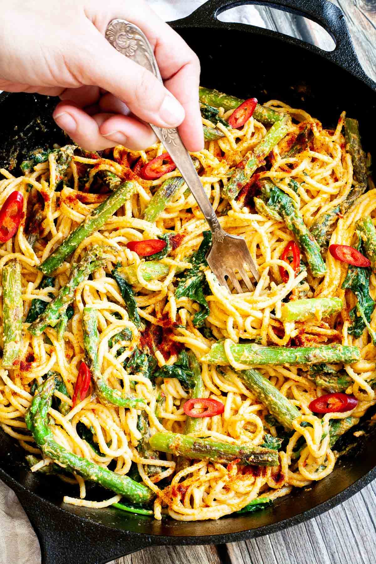 Black cast iron skillet with spaghetti in creamy light brown sauce with asparagus, spinach and red pepper slices. A hand is holding a fork to take a bite from the middle