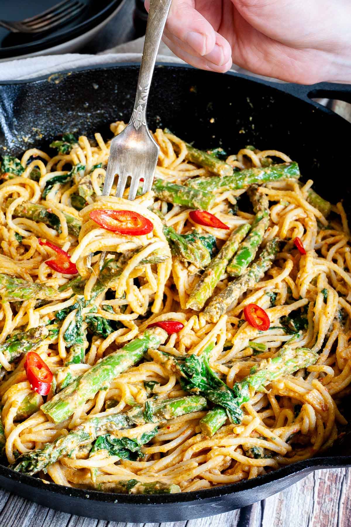 Black cast iron skillet with spaghetti in creamy light brown sauce with asparagus, spinach and red pepper slices. A fork is taking a bite from the middle.