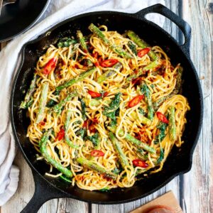 Black cast iron skillet with spaghetti in creamy light brown sauce with asparagus, spinach and red pepper slices