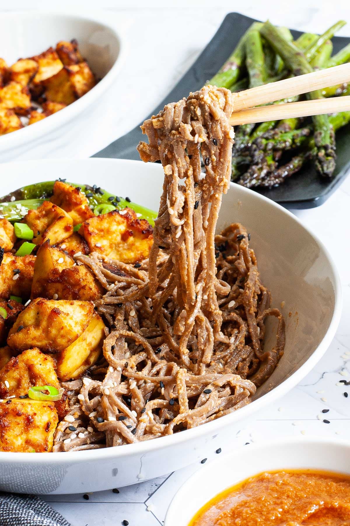 Dark brown noodles with crispy yellow crumbled tofu served in a white bowl. Chopsticks are lifting some of the noodles up. 