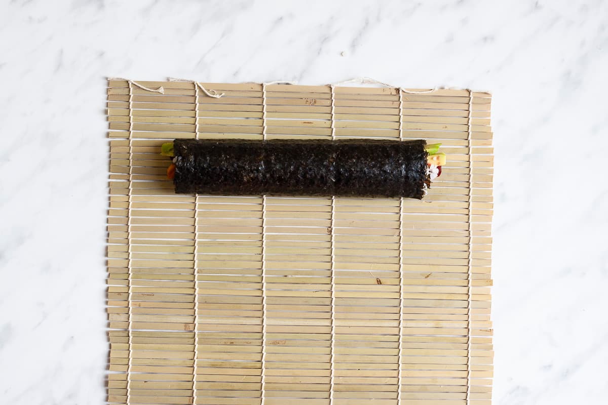 A bamboo mat with a long rolled sushi covered in seaweed sheets. Some of the fillings can be seen at the ends.