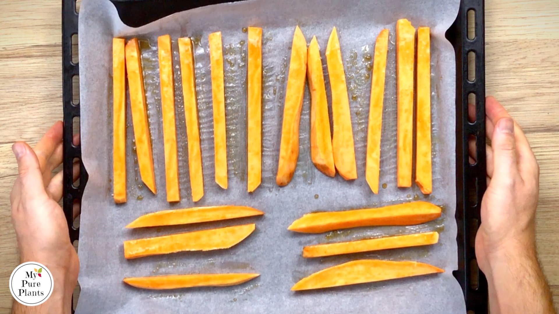 Sweet potato sticks are arranged in one layer on a parchment paper. A hand is holding the sheet pan to take it away for roasting.