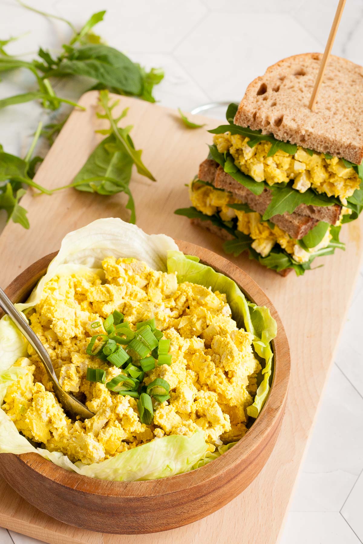 A wooden bowl with lettuce leaves topped with yellow egg salad. A stack of egg salad sandwich is right next to it.
