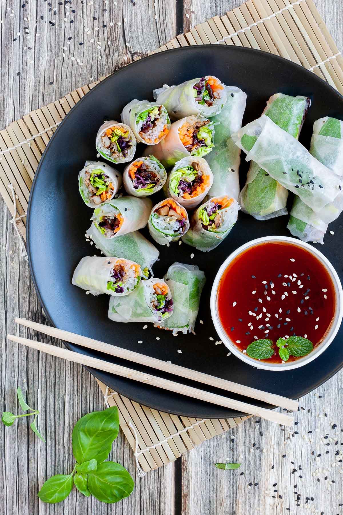 Opaque rice paper rolls filled with carrots, purple sprouts, thin glass. noodles and brown mushrooms are served on a large black plate. Several rolls are cut half so you can see the filling. Red sauce in a small white bowl is served next to it.