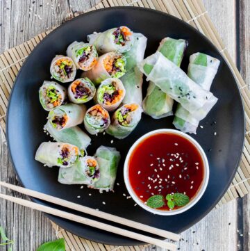 Opaque rice paper rolls filled with carrots, purple sprouts, thin glass. noodles and brown mushrooms are served on a large black plate. Several rolls are cut half so you can see the filling. Red sauce in a small white bowl is served next to it.