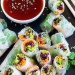 Opaque rice paper rolls filled with carrots, purple sprouts, thin glass. noodles and brown mushrooms are served on a large black plate. Several rolls are cut half so you can see the filling. Red sauce in a small white bowl is served next to it.