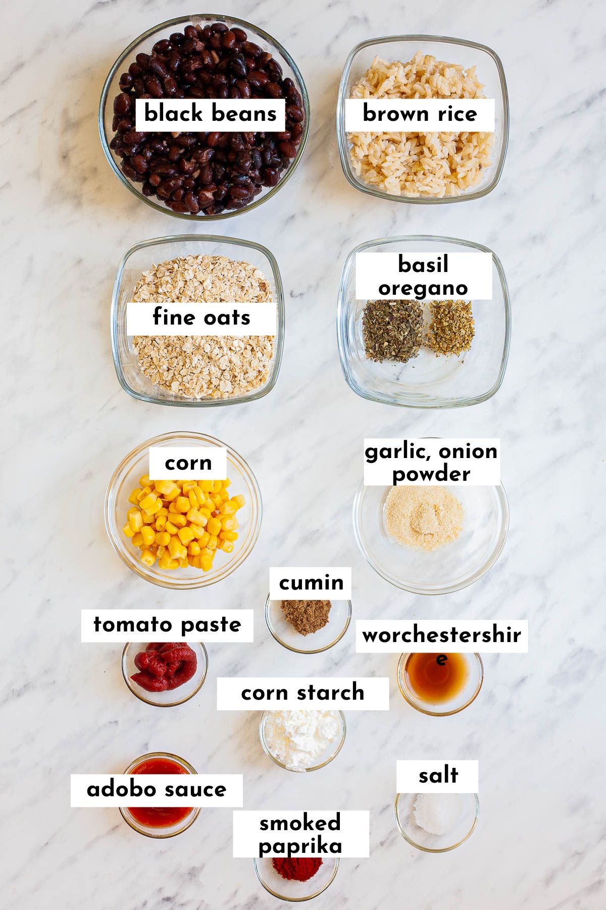 The ingredients of this black bean chipotle burger is displayed in small glass bowls: black bean, rice, oats, basil, oregano, corn, tomato paste and other spices