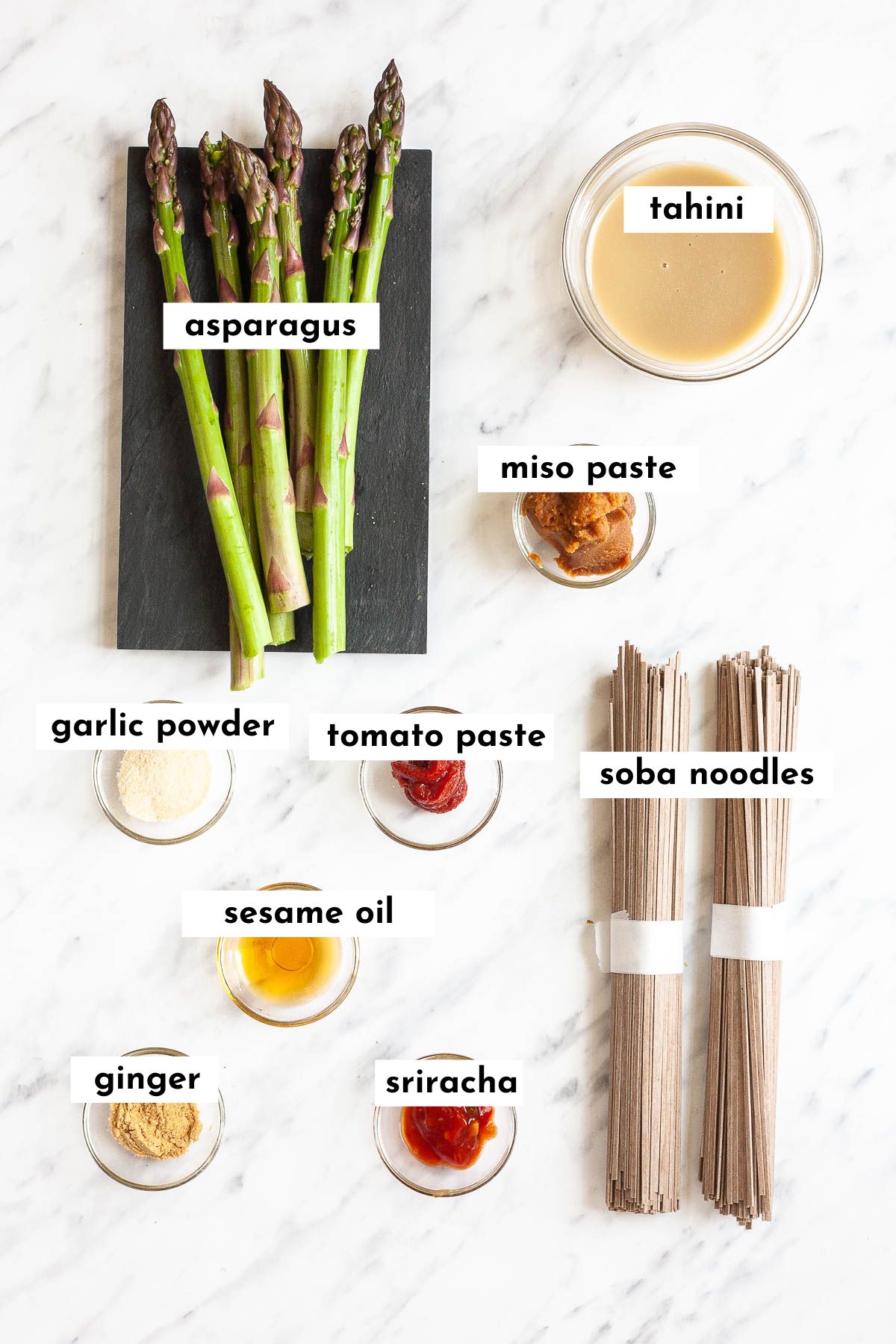 The ingredients of miso noodles is displayed including soba noodles, asparagus, and small glass bowls of miso paste, tahini, garlic powder, sesame oil, sriracha, tomato paste and ginger powder