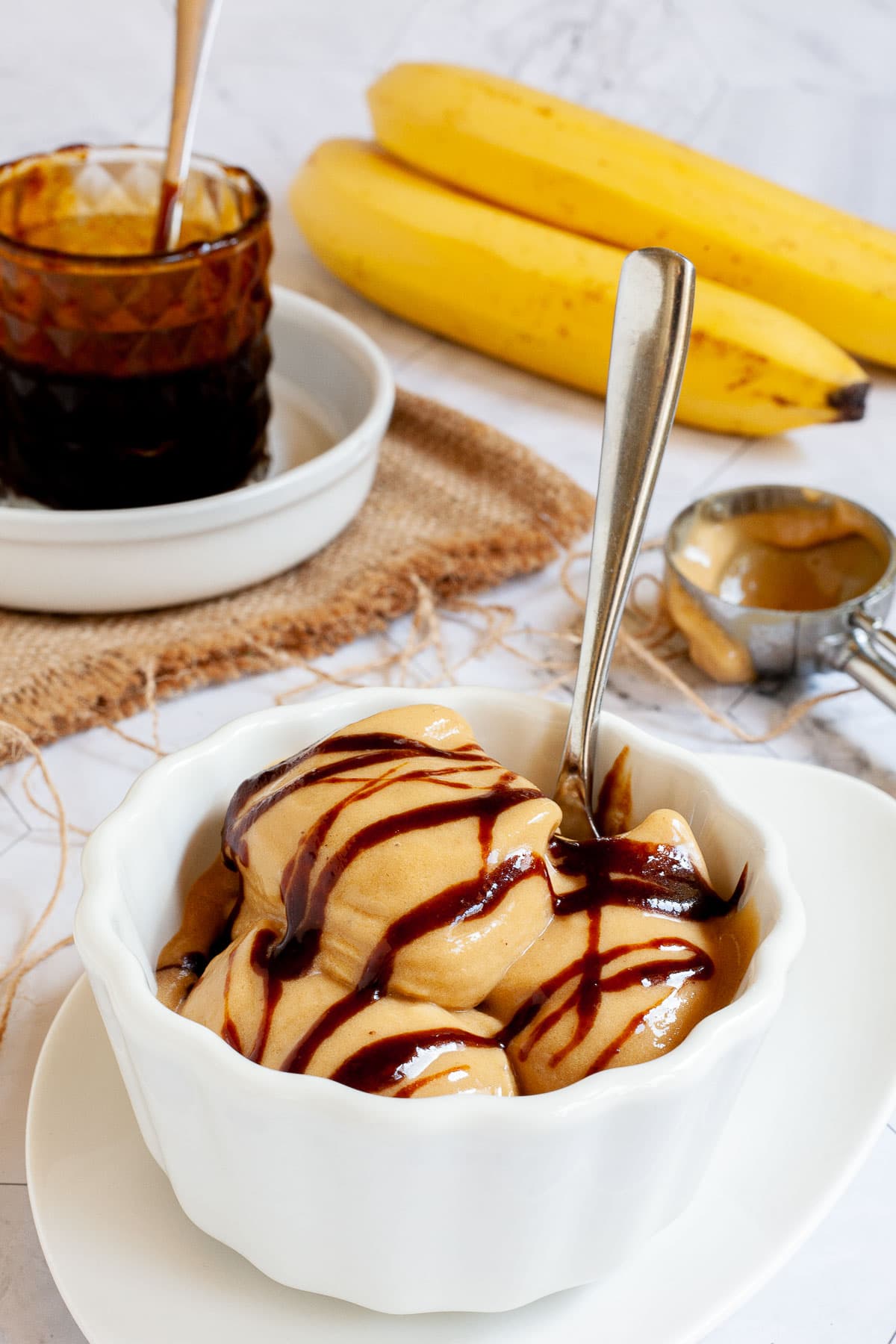 A white bowl of caramel colored ice cream with dark brown caramel drizzle on top. A glass jar of caramel sauce and 2 bananas are in the background. 