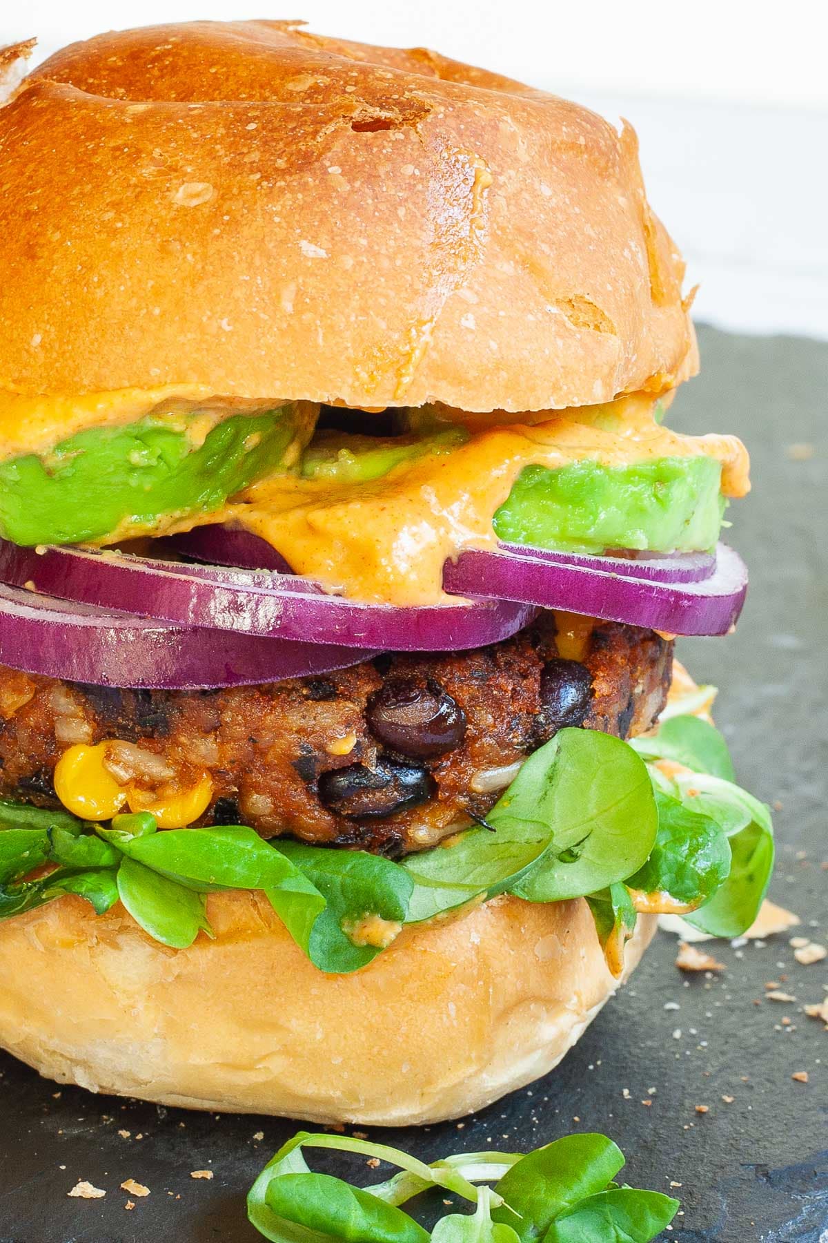 A burger bun with yellow sauce and green leaves is topped with a thick brown burger patty with visible sweet corn and black bean pieces, then red onion and avocado slices drizzled with yellow sauce again. It is covered with a top bun.