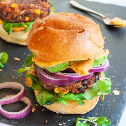 A burger bun with yellow sauce and green leaves is topped with a thick brown burger patty with visible sweet corn and black bean pieces, then red onion and avocado slices drizzled with yellow sauce again. It is covered with a top bun.
