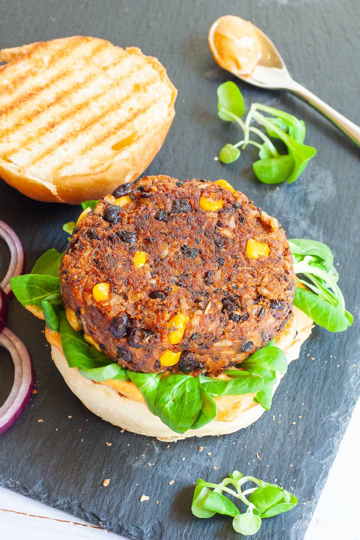 A burger bun with yellow sauce and green leaves is topped with a thick brown burger patty with visible sweet corn and black bean pieces. The top bun is next to it.