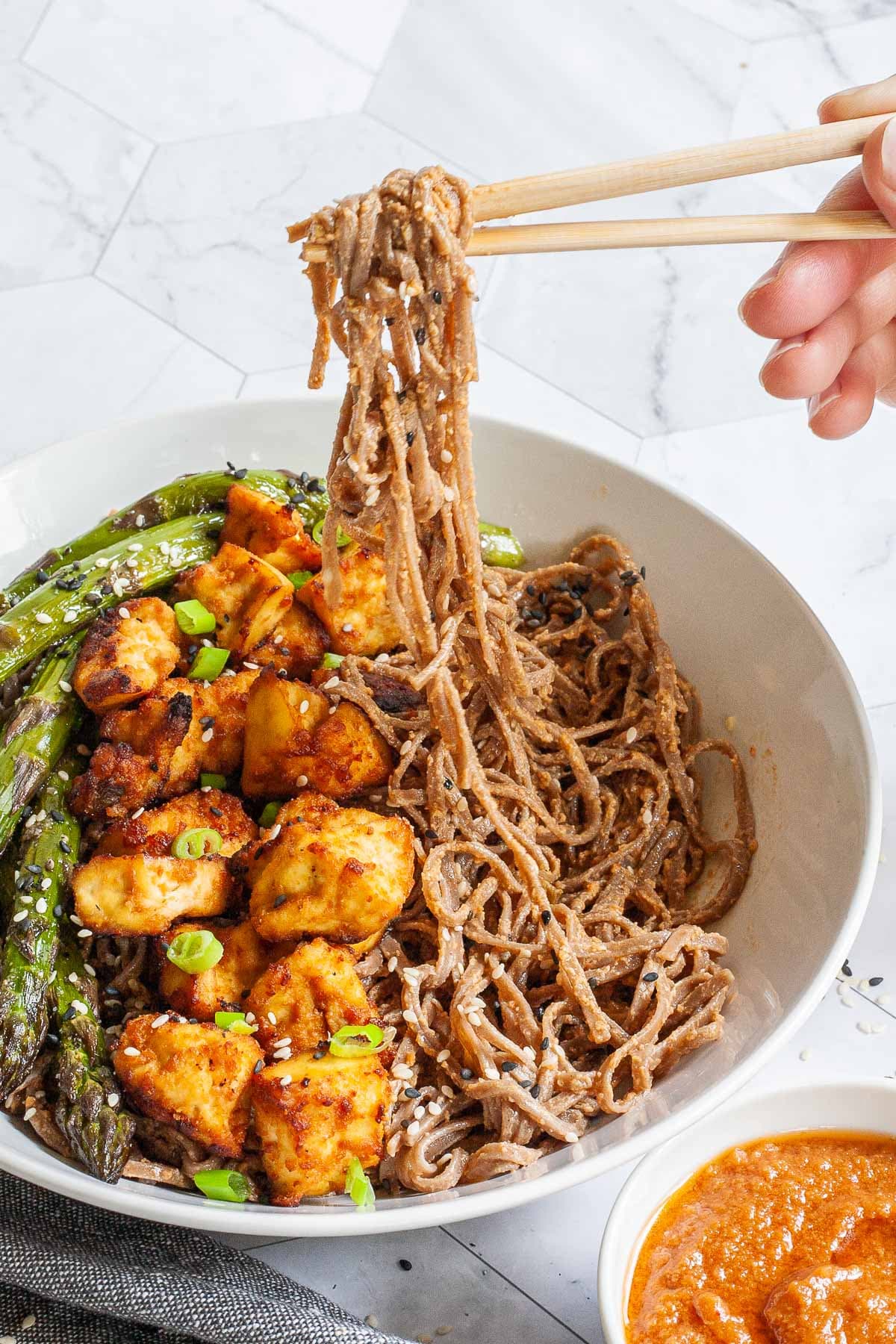 Large white bowl with brown soba noodles, orange crispy tofu bites, roasted asparagus and black and white sesame seeds. A hand is holding chopsticks and lifting some noodles up.