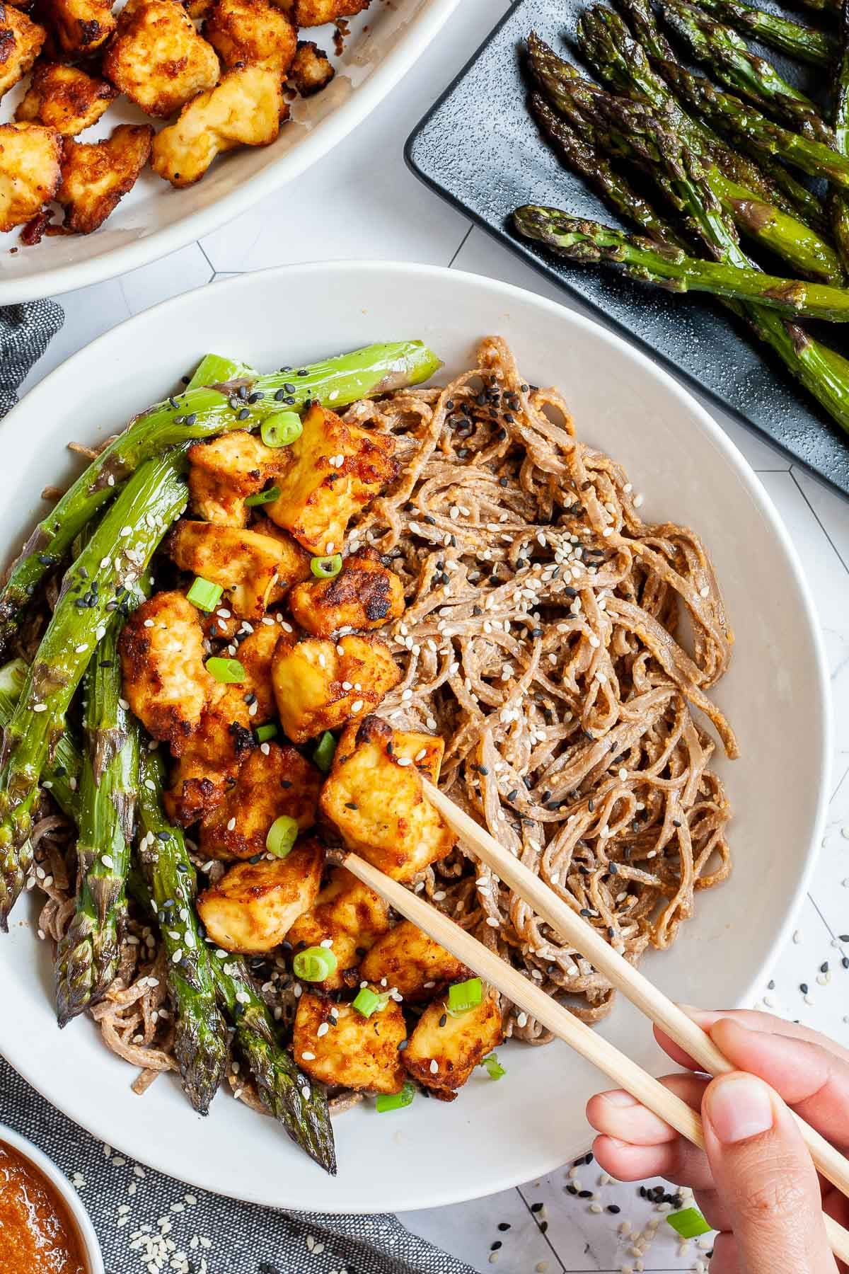 Large white bowl with brown soba noodles, orange crispy tofu bites, roasted asparagus and black and white sesame seeds. More asparagus and tofu bites are on the side. A hand is holding chopsticks and taking one tofu.