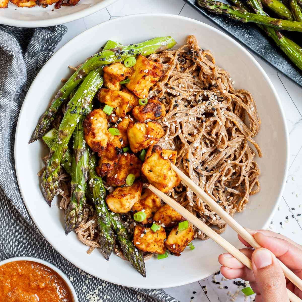 Large white bowl with brown soba noodles, orange crispy tofu bites, roasted asparagus and black and white sesame seeds. More asparagus and tofu bites are on the side. A hand is holding chopsticks and taking one tofu.