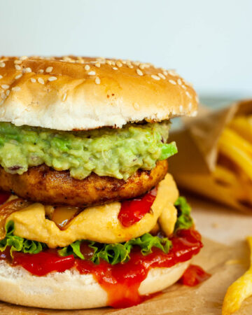 A burger with ketchup, lettuce, yellow cheese sauce, bell peppers, guacamole and grilled mushroom cap on brown paper. French fries are placed next to it.