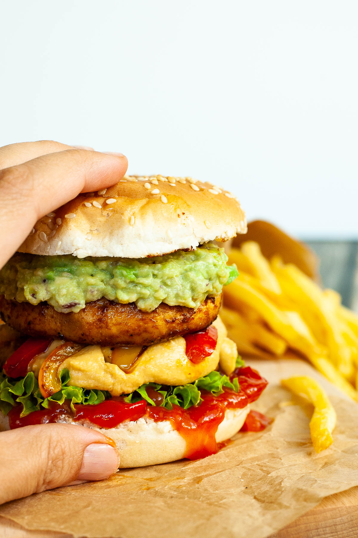 A hand is holding a burger with ketchup, lettuce, yellow cheese sauce, bell peppers, guacamole and grilled mushroom cap on brown paper. French fries are placed next to it.