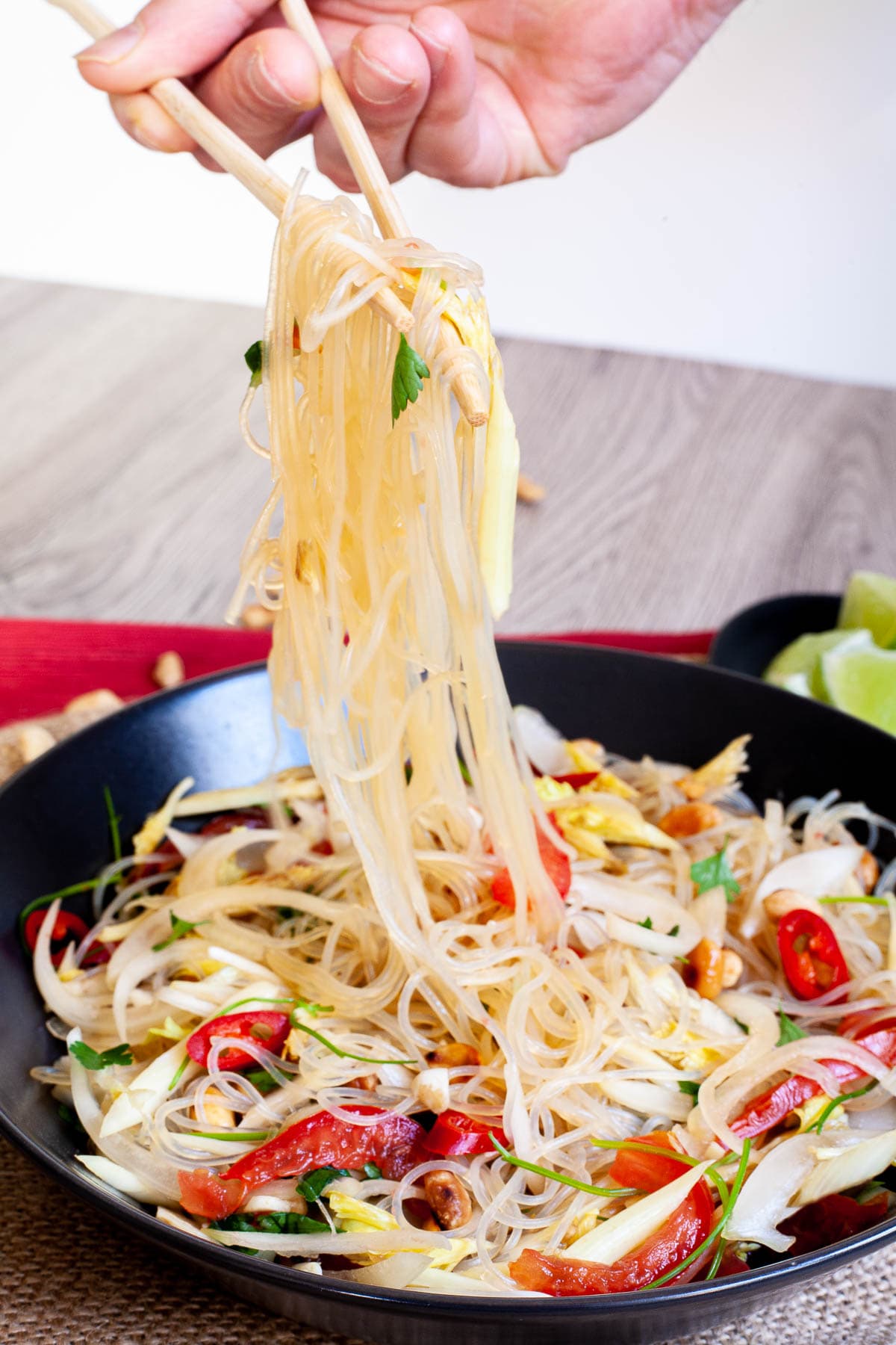 Black plate on a red mate with thin glass noodles, sliced tomatoes, roasted peanuts, sliced onion, sliced chili peppers, chopped green herbs. A hand is holding chopsticks and lifting thin noodles up the air.