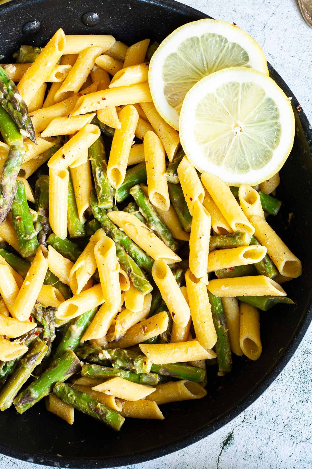 A frying pan with penne pasta, asparagus pieces and 2 lemon slices. 