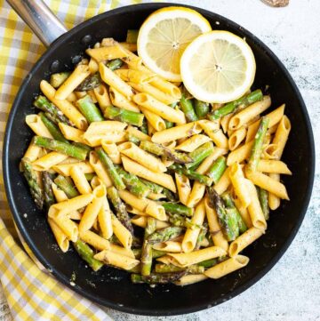 A frying pan with penne pasta, asparagus pieces and two lemon slices
