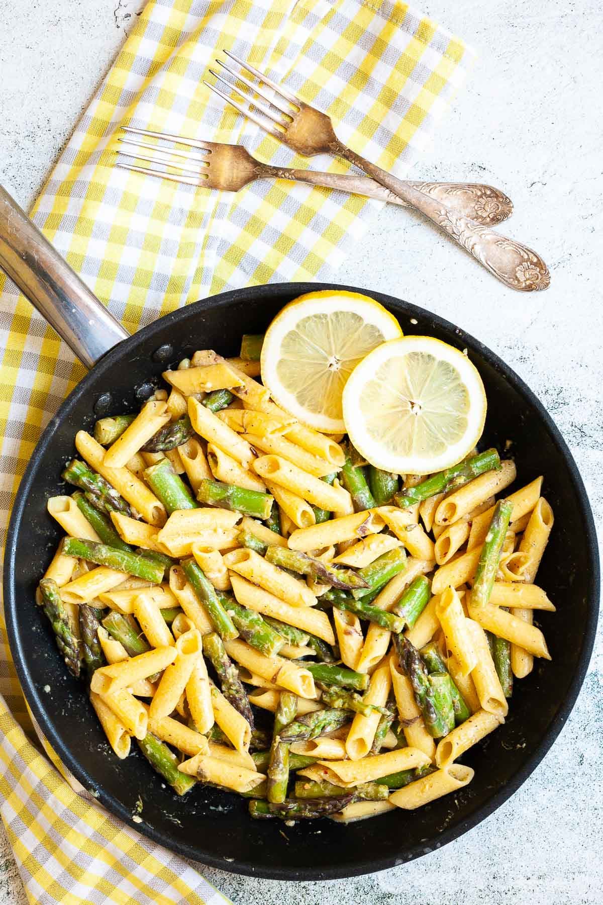 A frying pan penne pasta, asparagus pieces and two lemon slices. 
