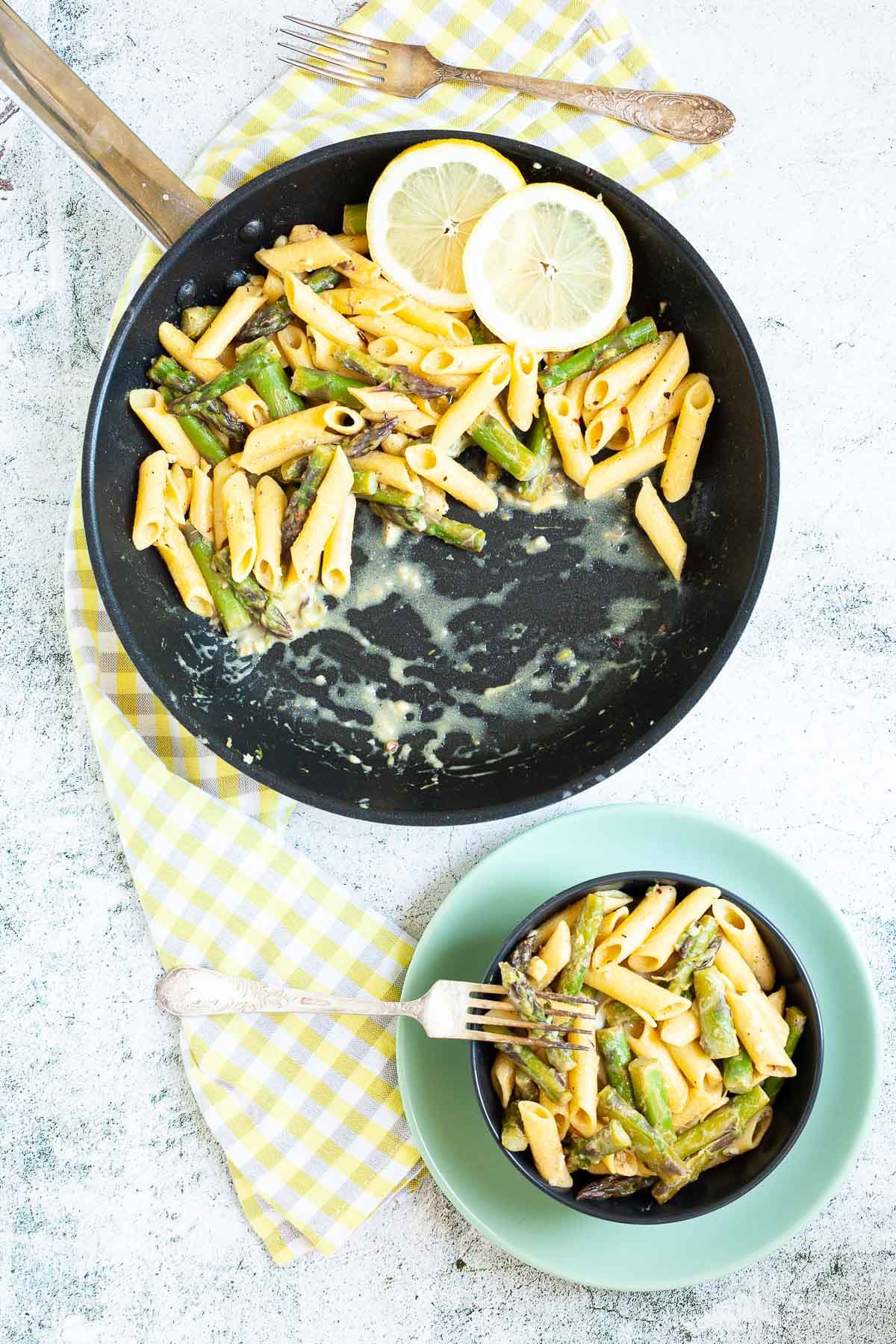 A frying pan and a black bowl next to it both with penne pasta and asparagus pieces. 