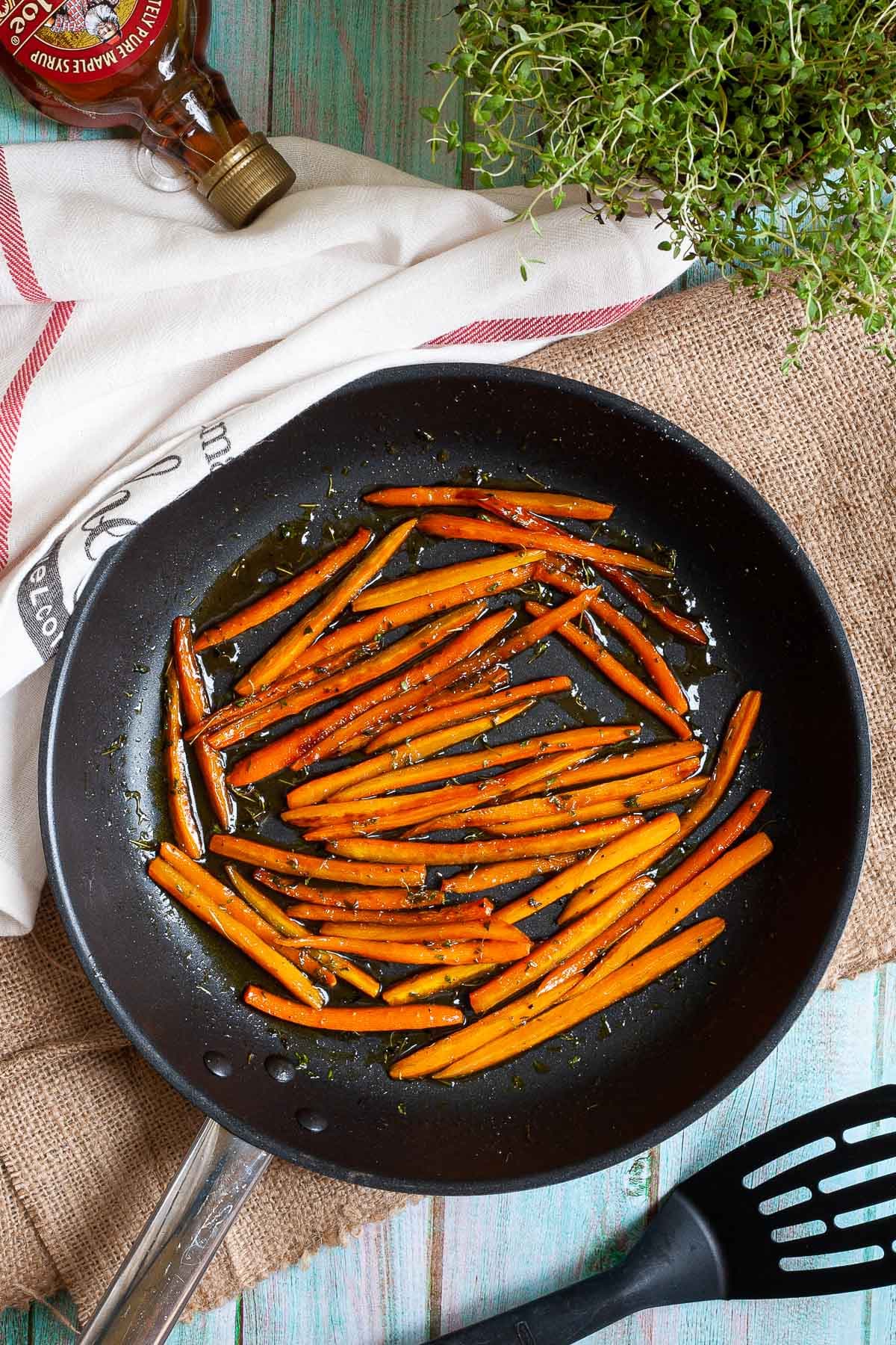Frying pan with matchstick carrots in a glaze. Maple syrup and fresh thyme is next to it. 