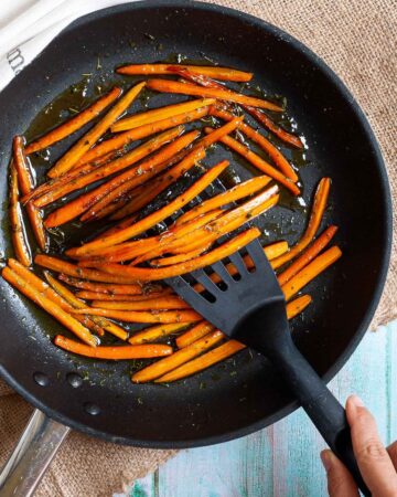 Frying pan with matchstick carrots in a glaze. A hand is holding a slotted turner and taking some from the pan.