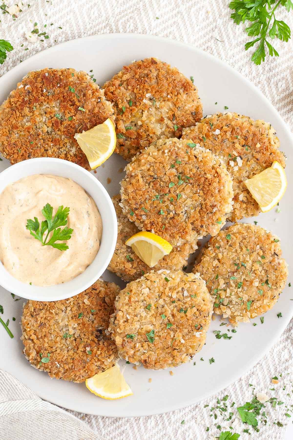 Several round-shaped breadcrumbs covered patties on a large white plate with a light orange sauce in a white bowl. Chopped parsley and lemon wedges are around for decoration. 